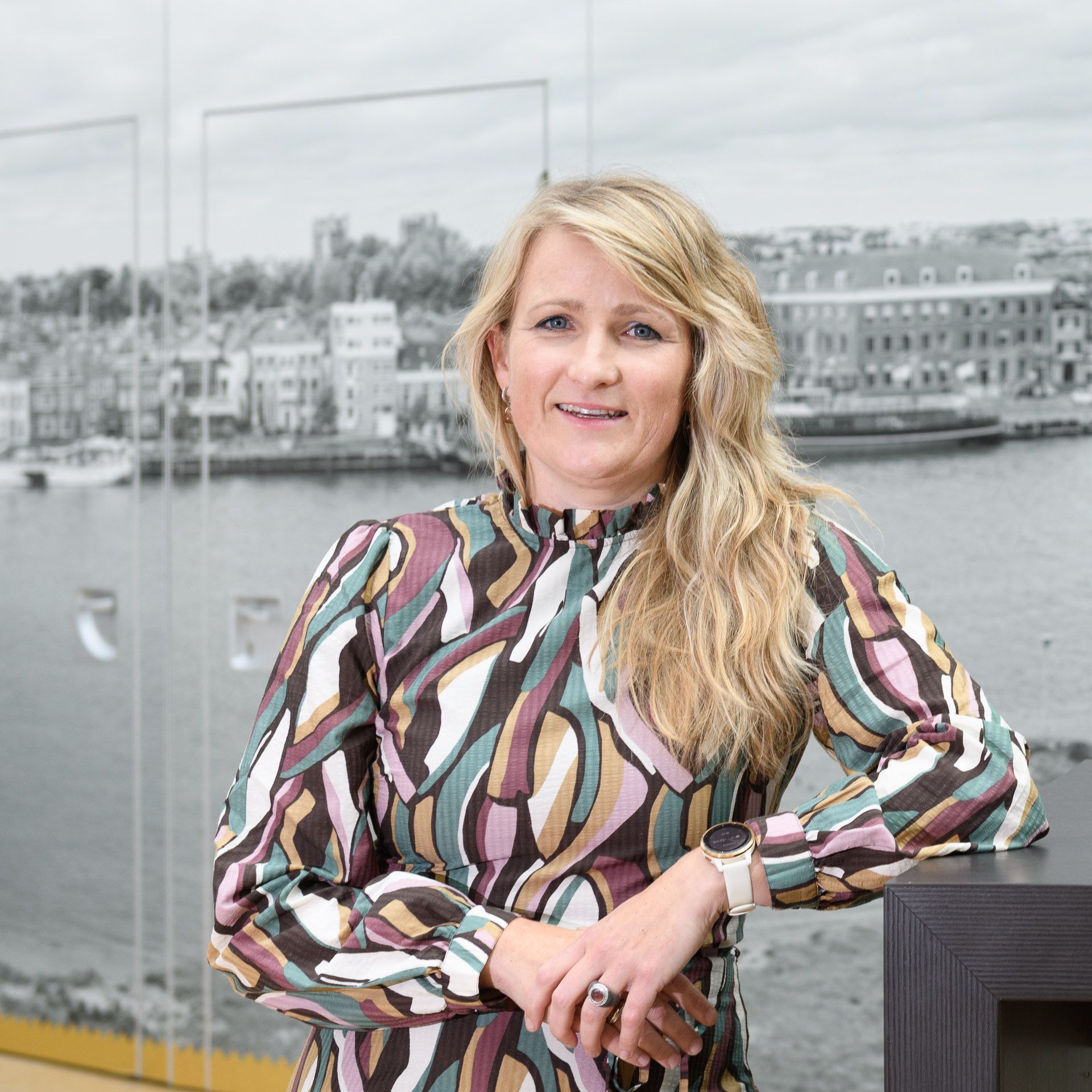 Een vrouw in een jurk met patroon leunt glimlachend tegen een toonbank, met op de achtergrond een havengezicht.