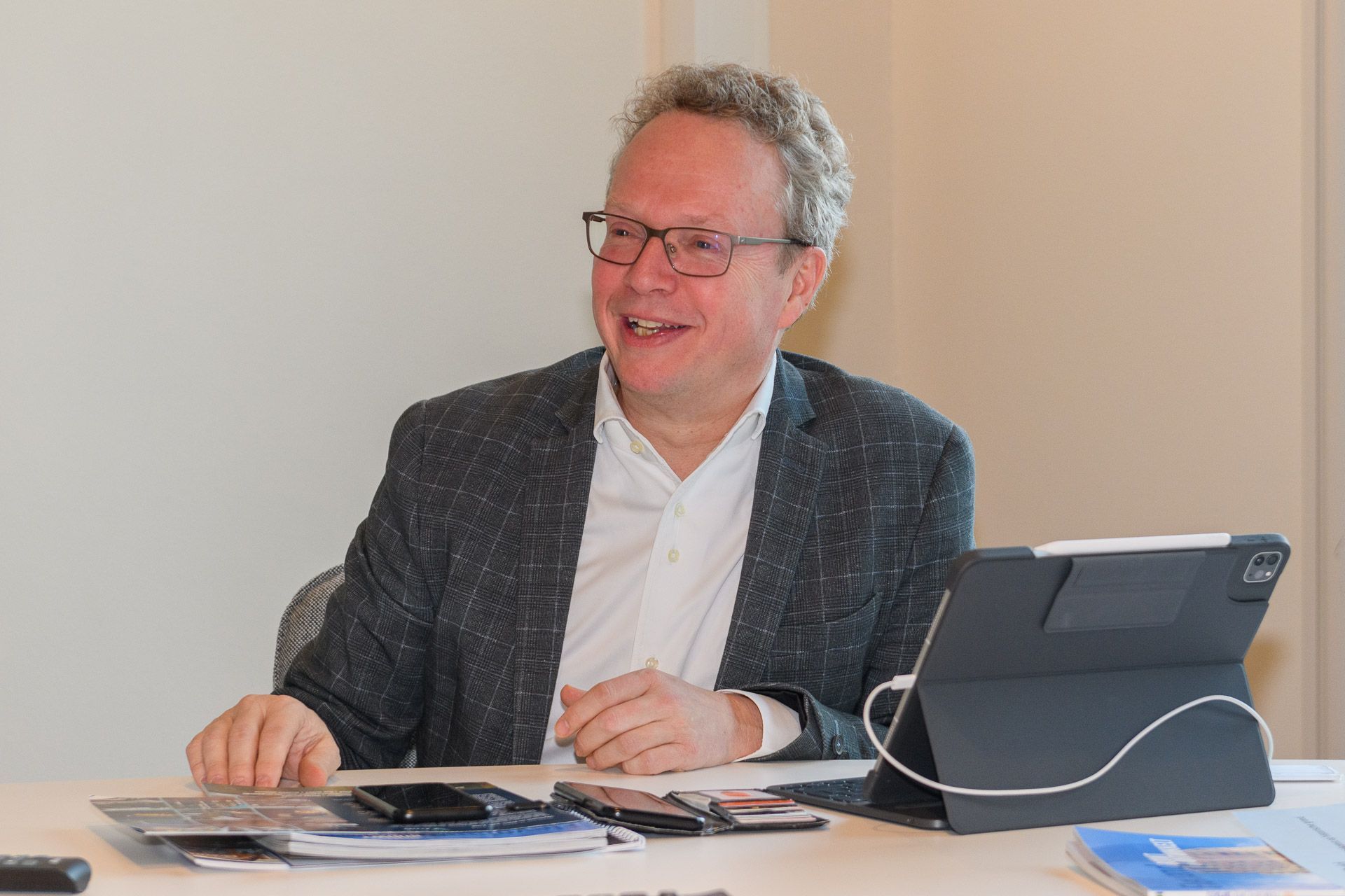Een man met een bril en een blazer zit glimlachend aan een tafel met elektronische apparaten.