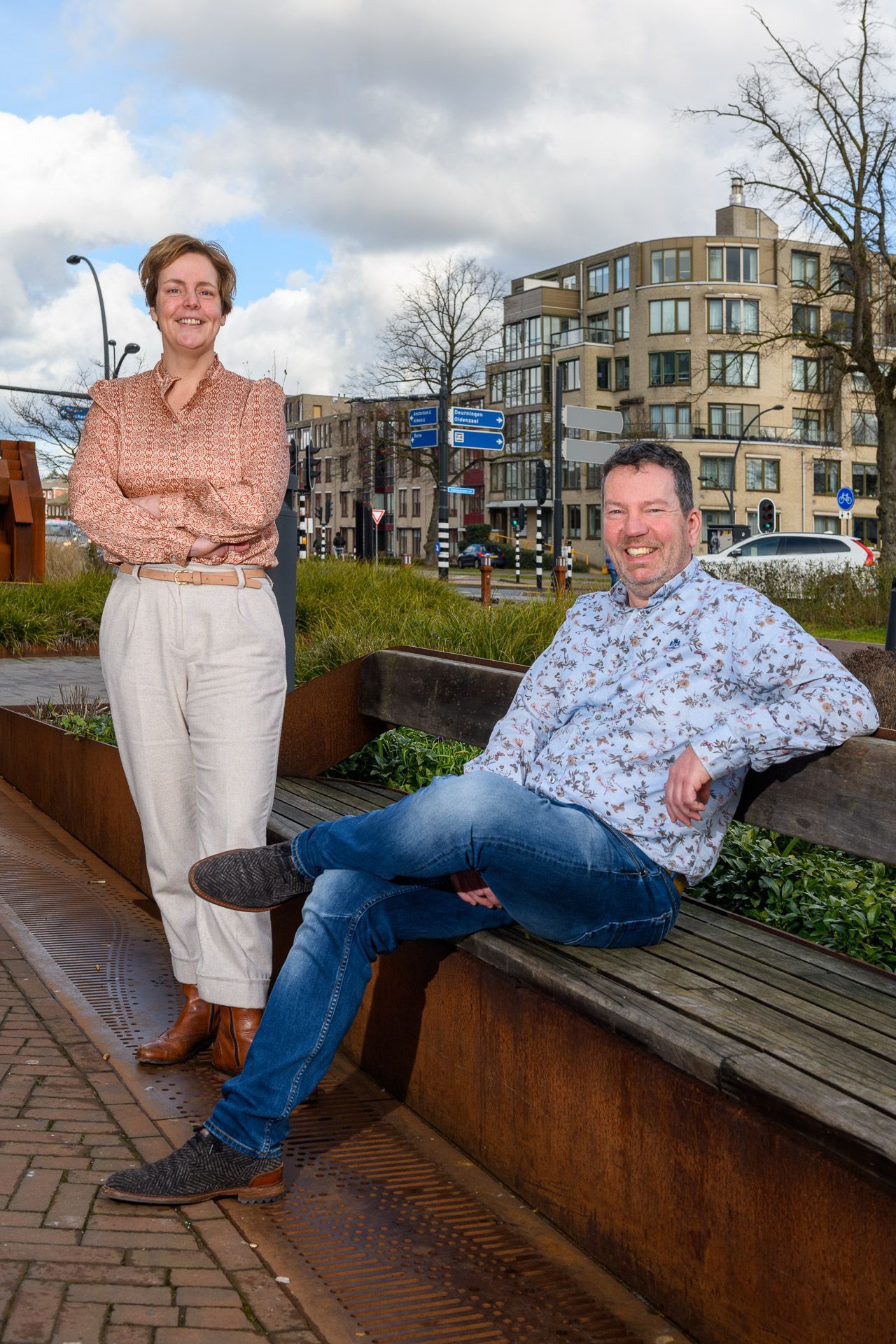 Een vrouw staat met haar armen over elkaar. Een man zit op een bankje en glimlacht. Moderne stedelijke omgeving, zonnige dag.