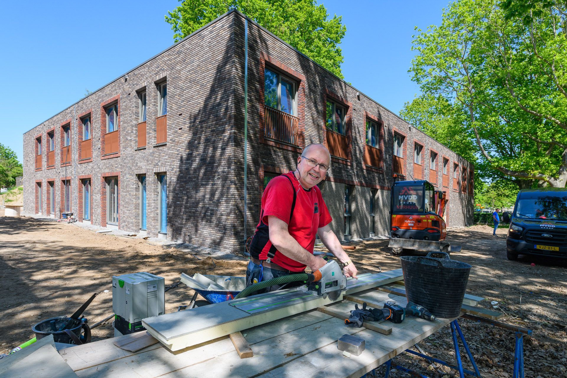 Een man snijdt een plank buiten een met hout bekleed gebouw. Bouwplaats.