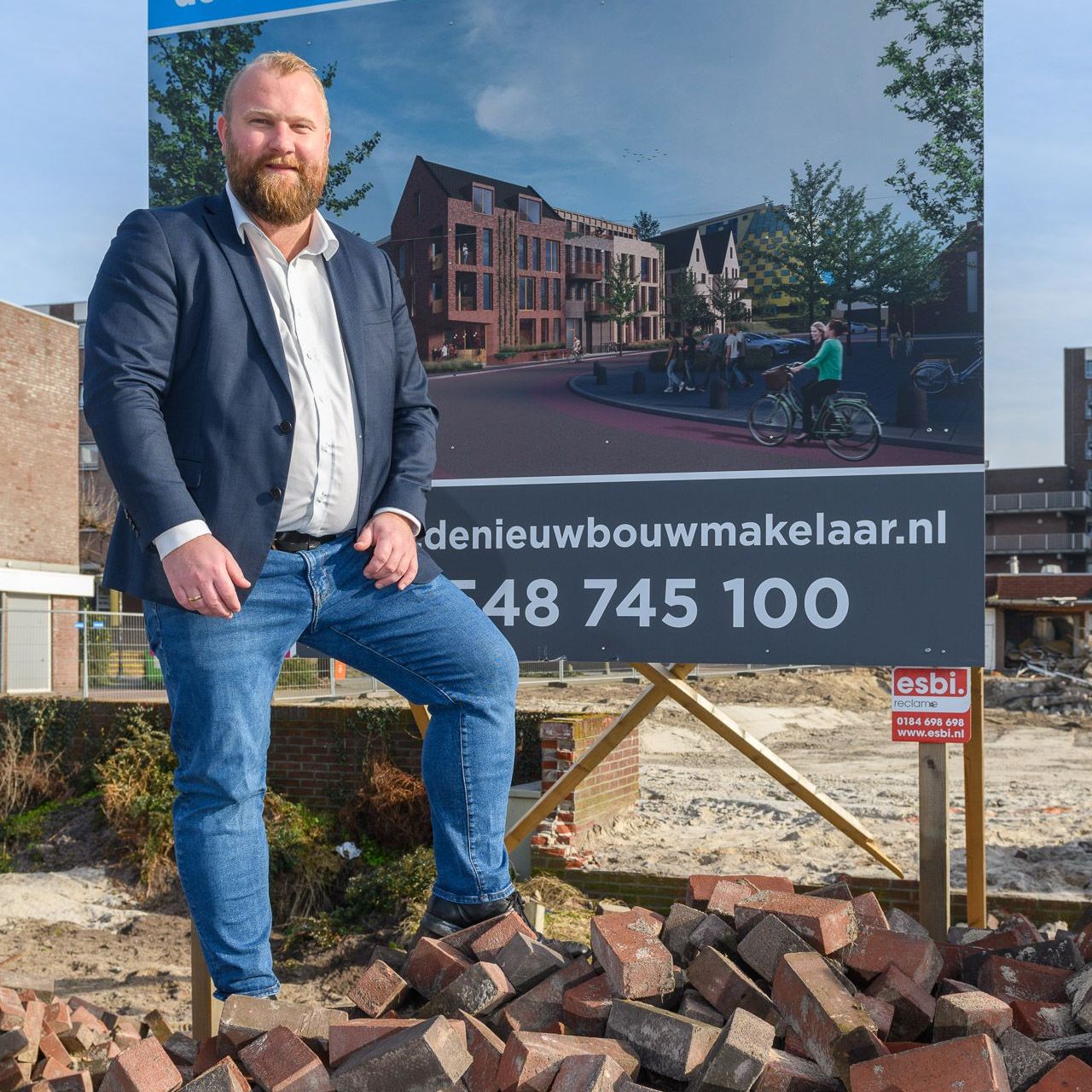 Een man in een blazer en spijkerbroek staat op bakstenen voor een makelaarsbord.