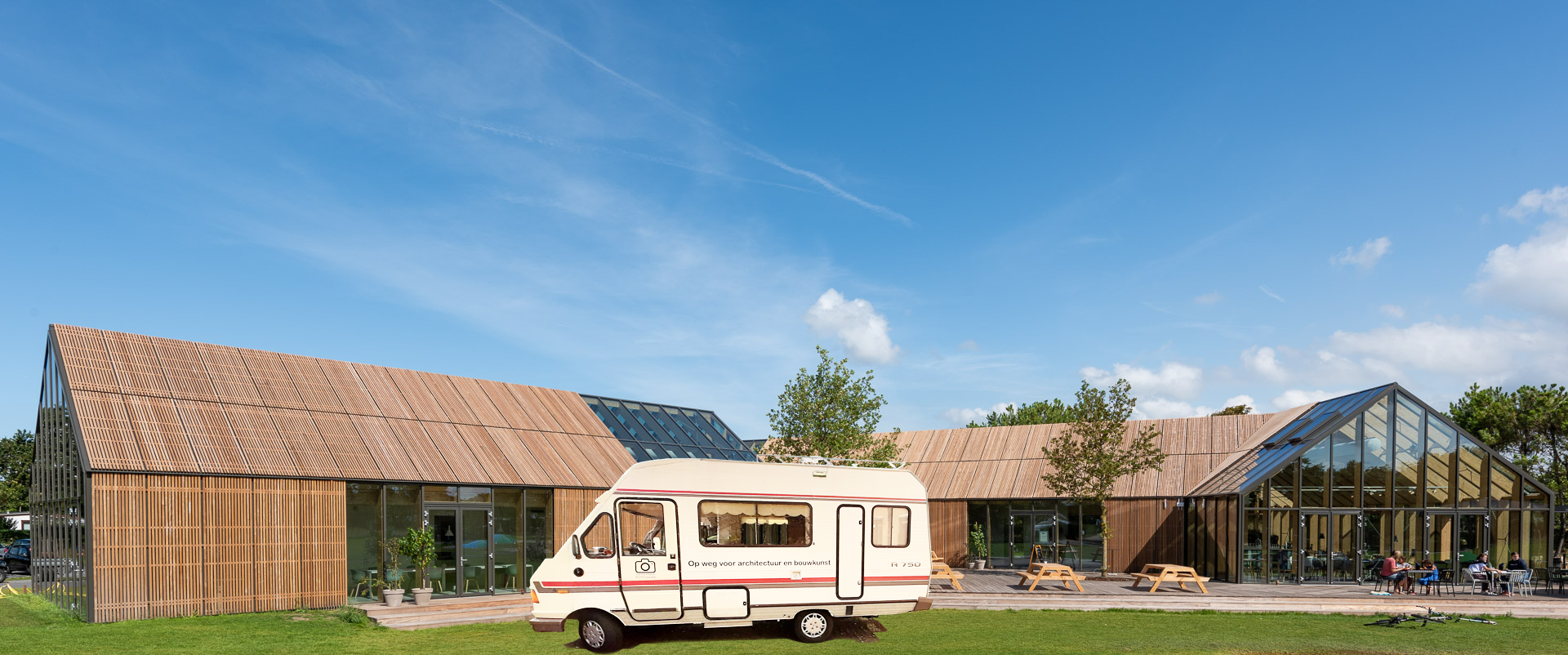 Een camper staat geparkeerd voor moderne houten gebouwen met grote ramen, onder een blauwe hemel.
