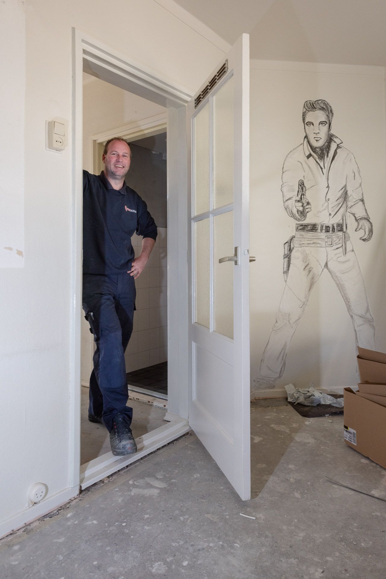 Man in de deuropening, glimlachend. Witte deur en kozijn. Muurschildering van Elvis op de muur.