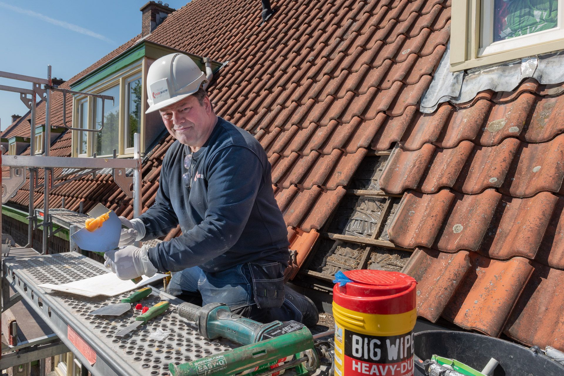 Een dakdekker repareert een pannendak. Hij knielt op een steigerplatform en houdt gereedschap en doekjes vast in de zon.