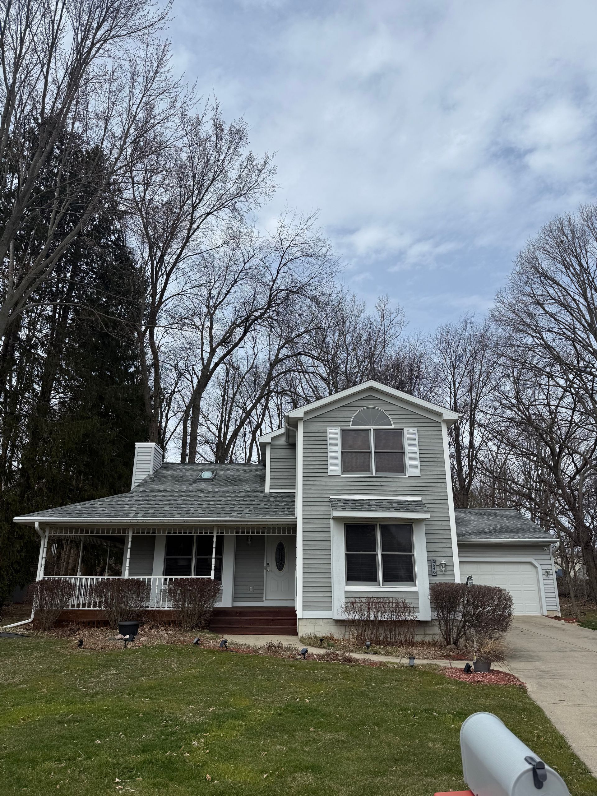 Two-story white house with a front porch, gray roof, and leafless trees in a cloudy yard.