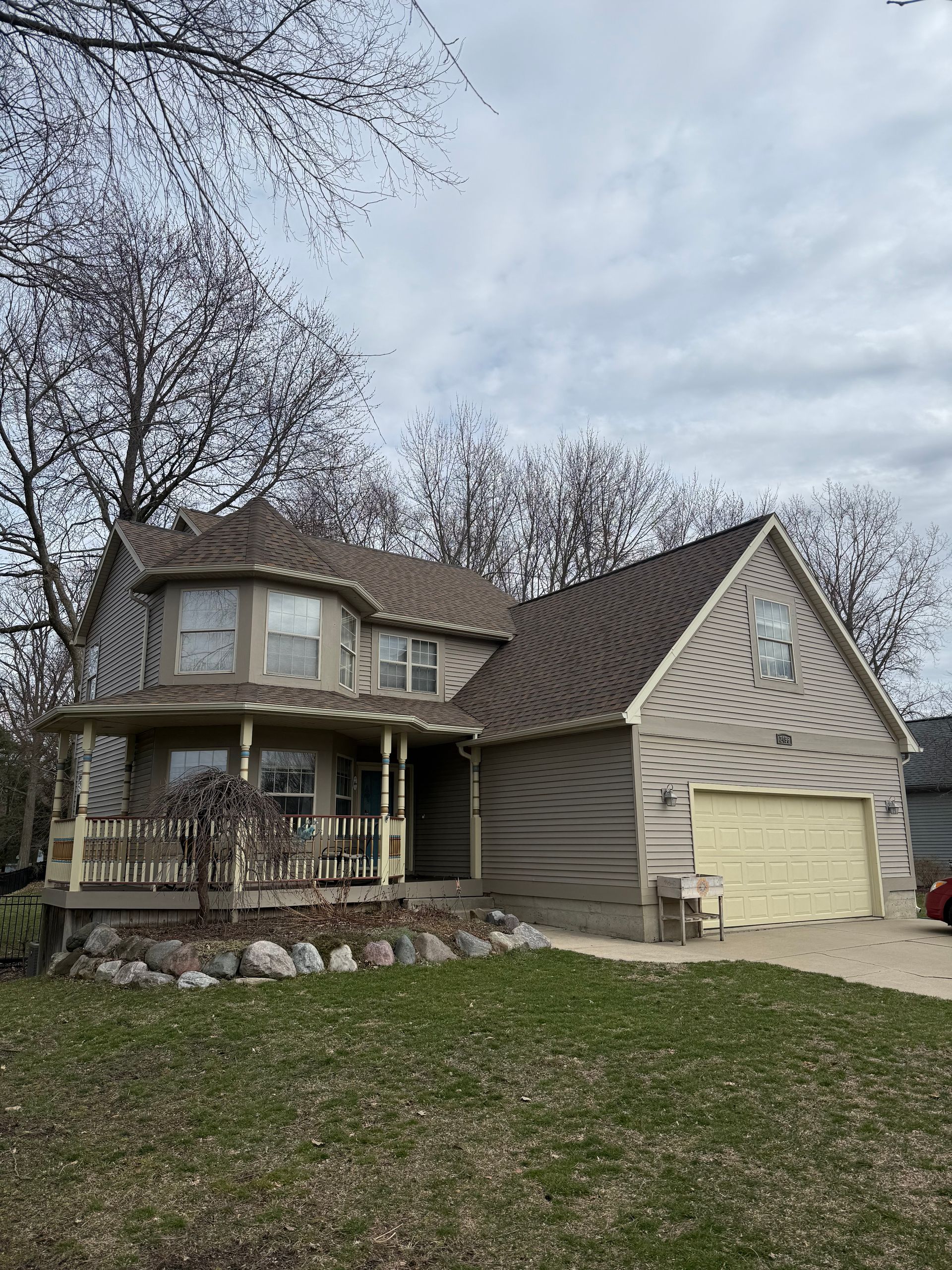 Suburban two-story beige house with a front porch, attached garage, and leaf-covered lawn under an overcast sky