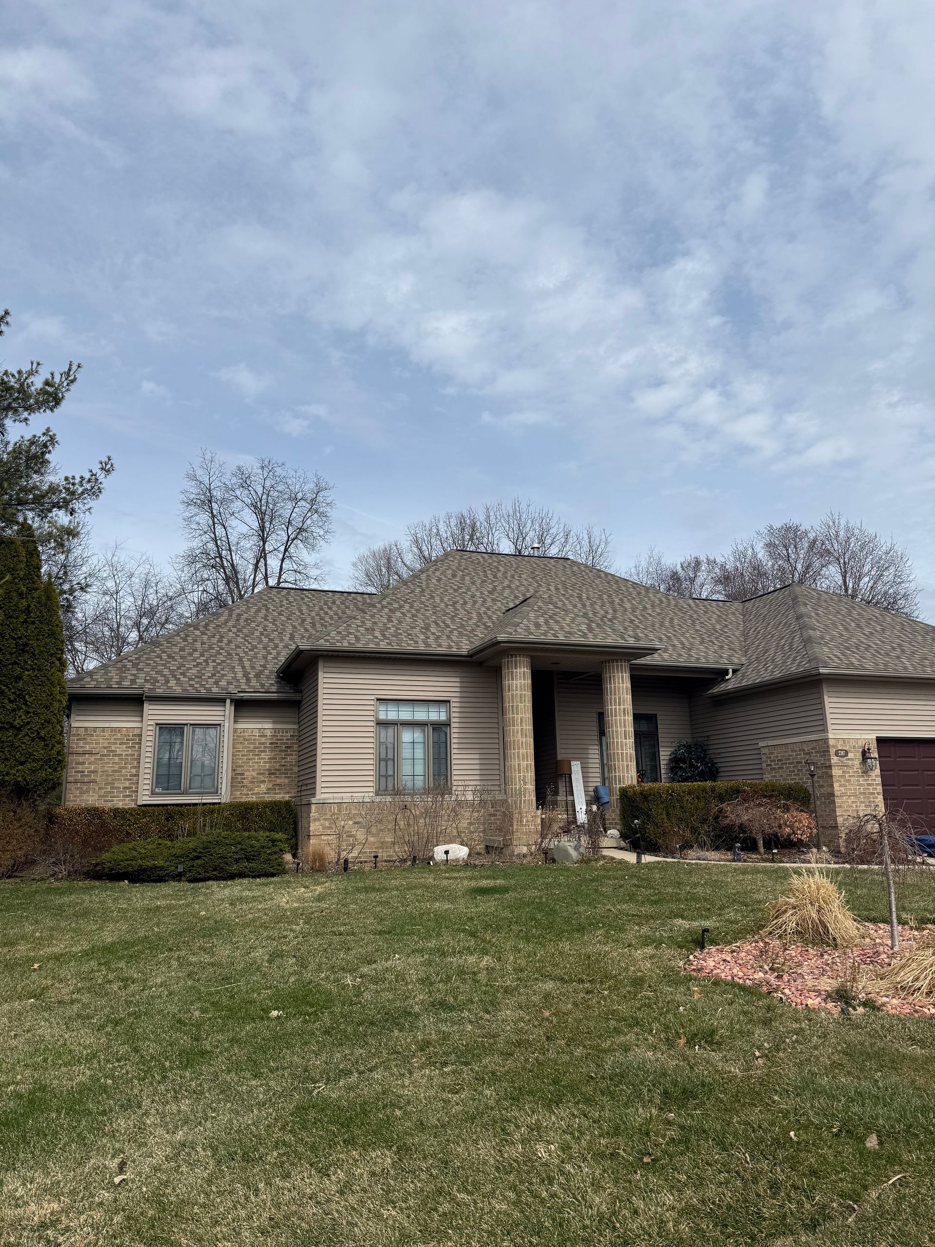 Single-story stone house with a shingled roof and front lawn under a partly cloudy sky