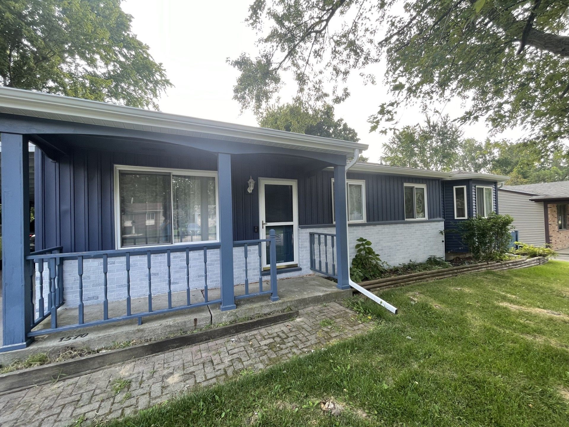 Blue single-story house with a small front porch, railing, and grassy yard under trees