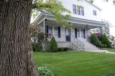 White two-story house with front porch, green lawn, and a large tree in the foreground.