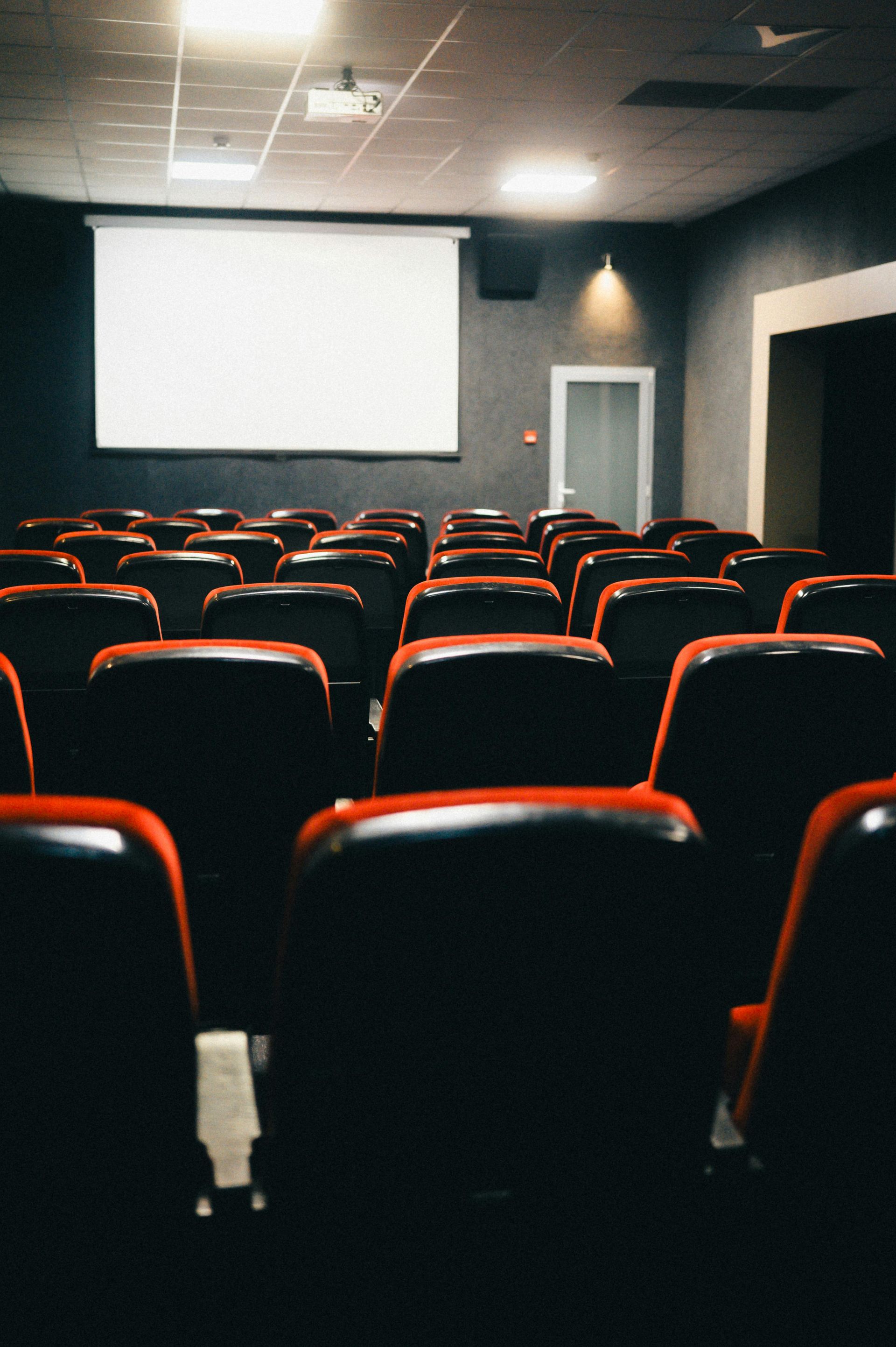 Empty cinema with red chairs and a blank screen, dimly lit.