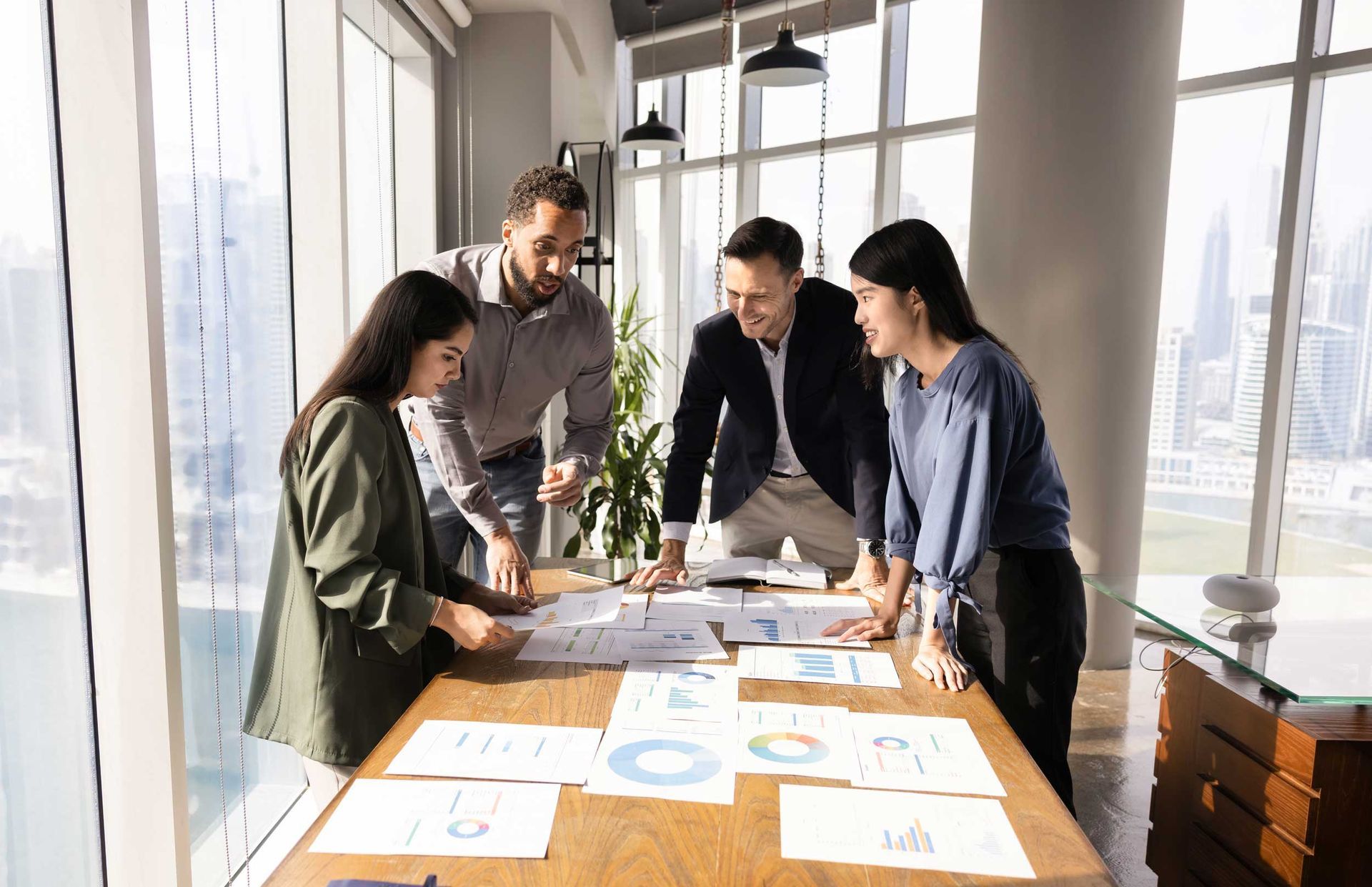Four coworkers reviewing charts and documents around a sunlit conference table in an office.