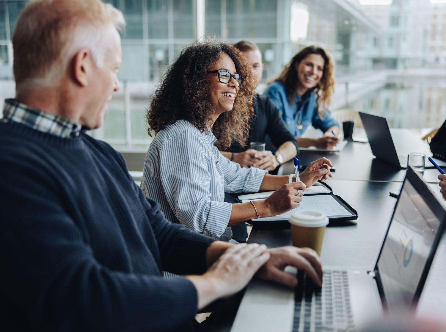 Colleagues in a bright conference room, smiling and talking around a meeting table with laptops and coffee cups