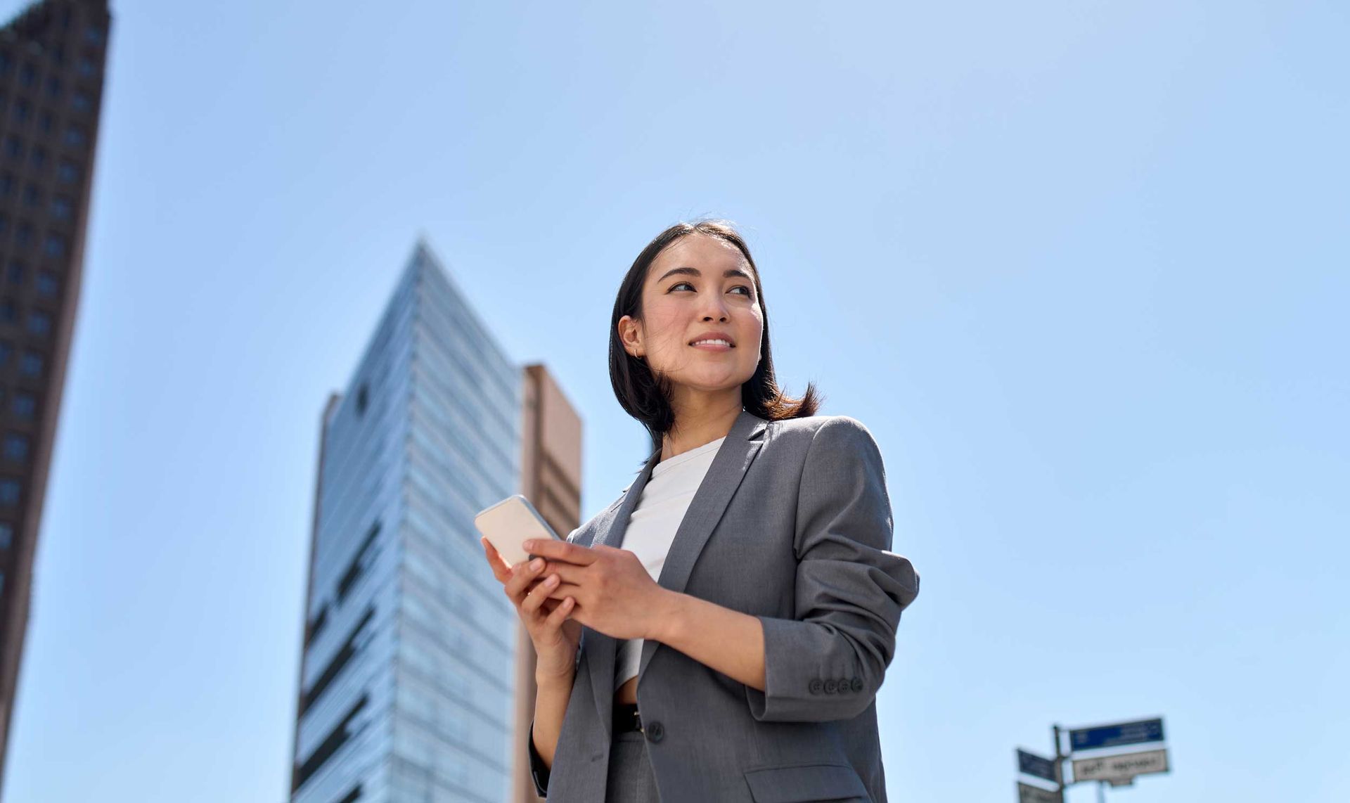 Businessperson in a gray suit looking up beside modern office buildings under a blue sky