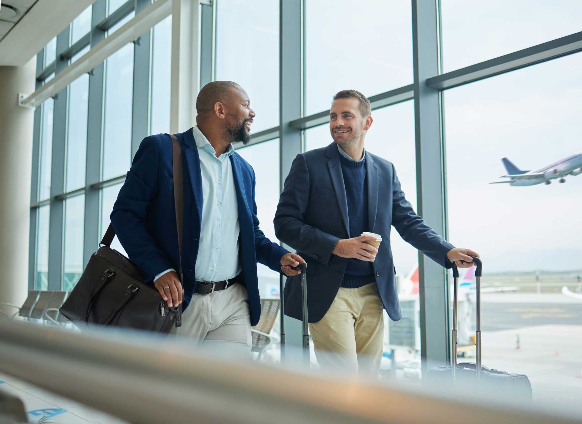 Two travelers in a bright airport terminal walk with luggage, chatting by large windows.