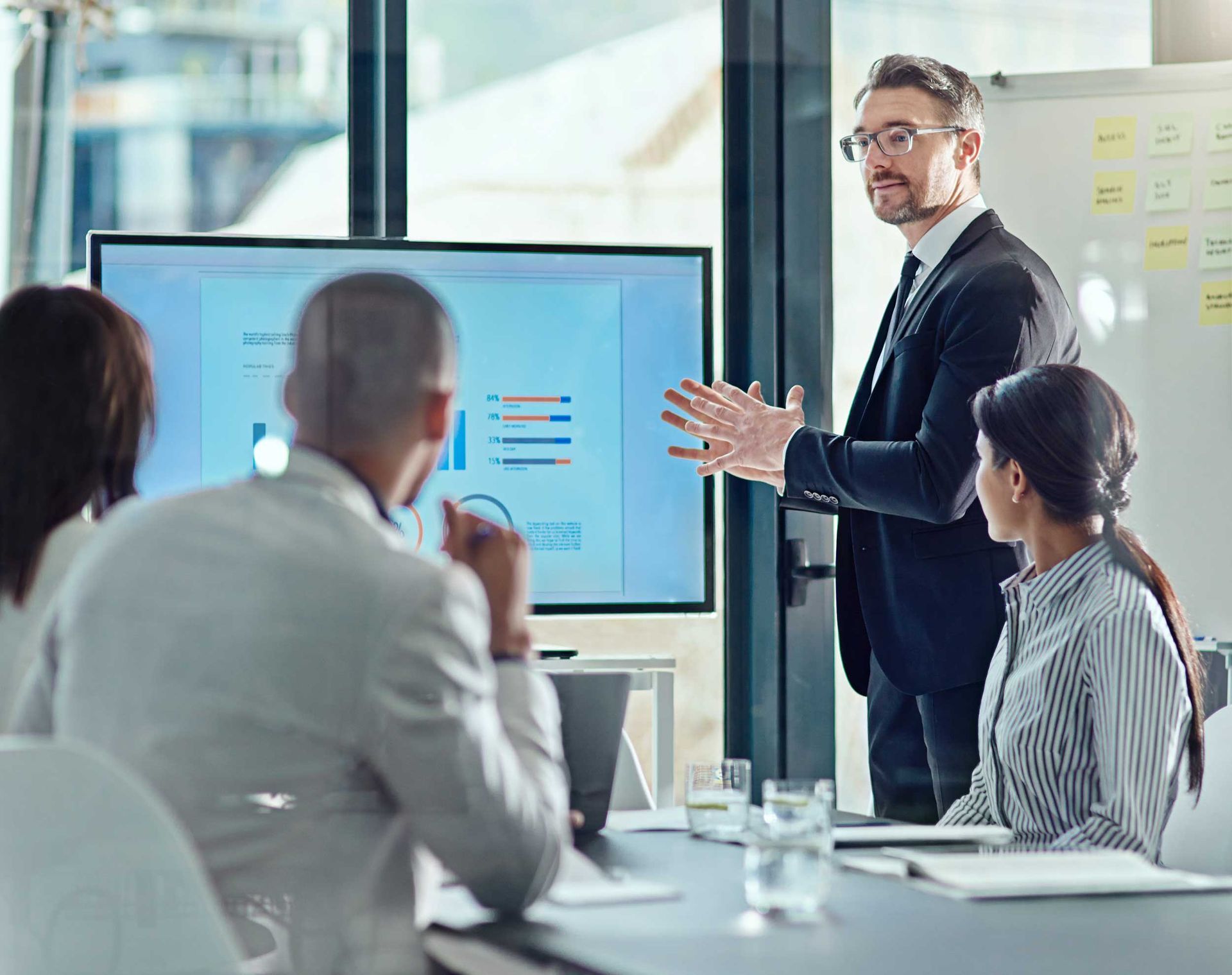Presenter discussing charts on a monitor during a business meeting, with two colleagues listening.
