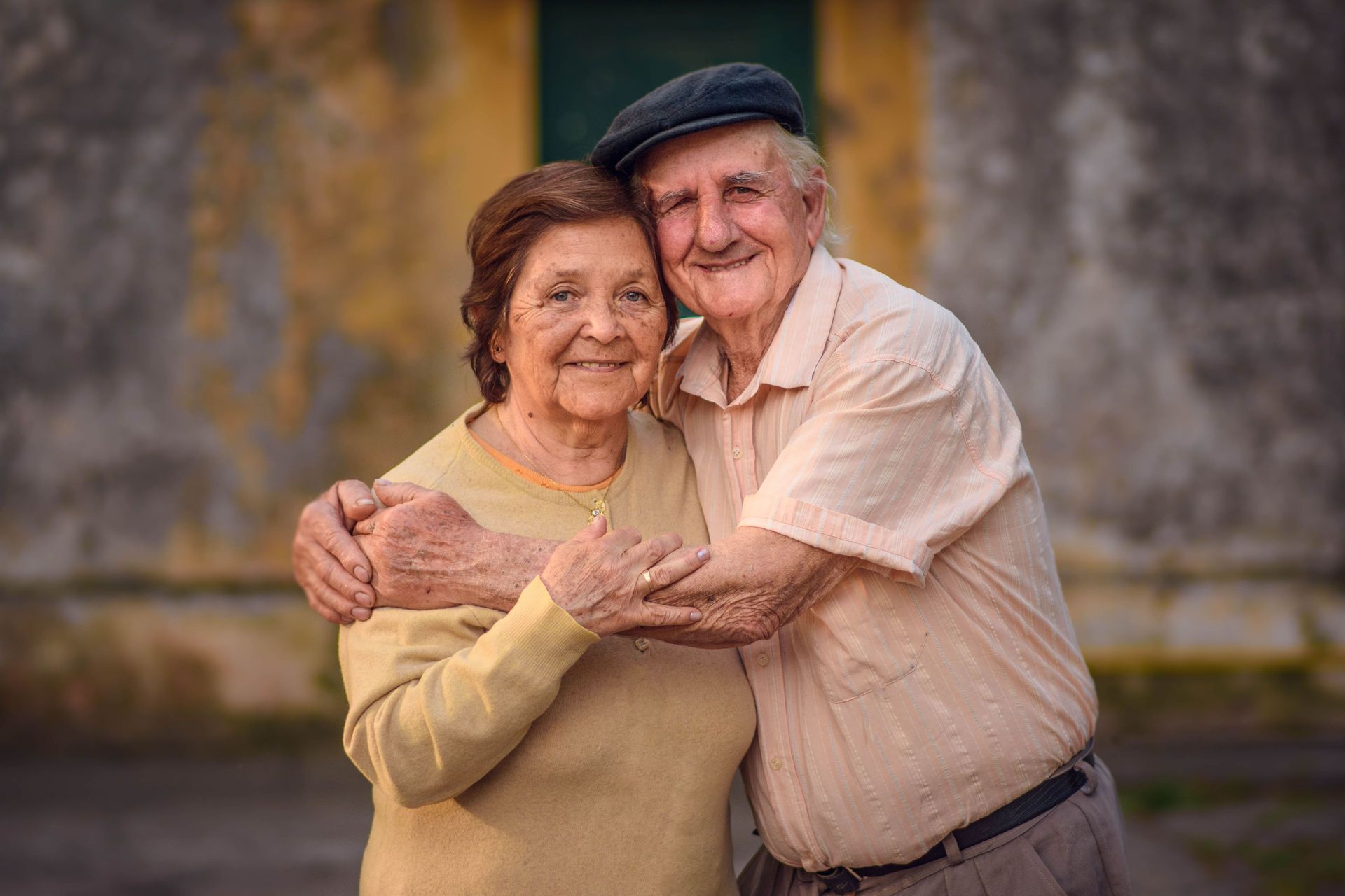 Smiling elderly couple hugging outdoors against a blurred wall background