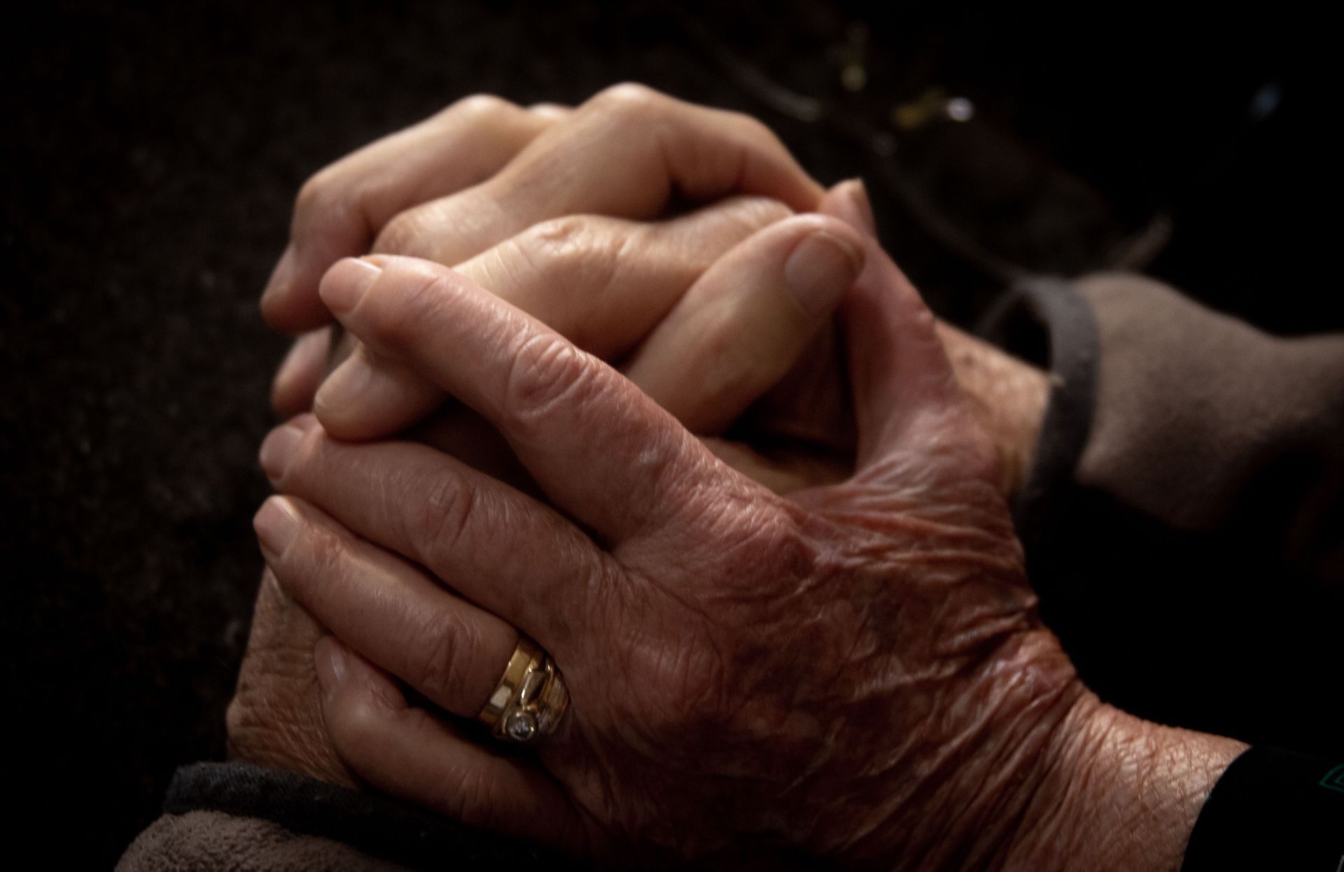 Two hands clasped together, one wearing a gold ring, against a dark background