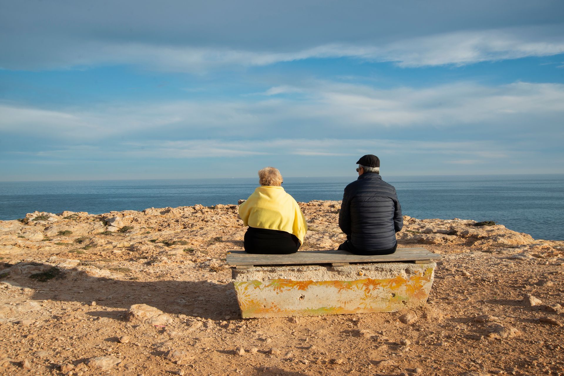 Two people sit on a bench facing the ocean under a blue sky.