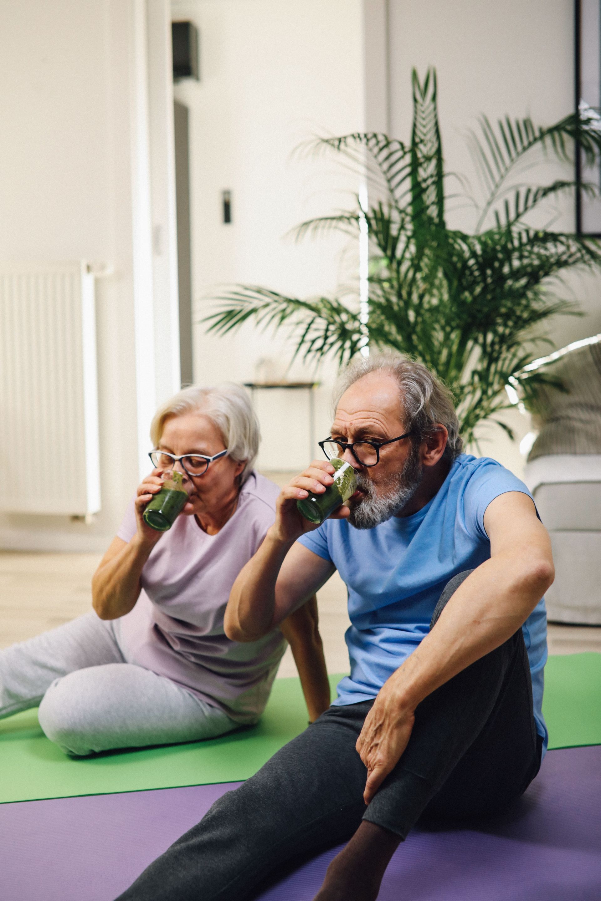 Two people doing seated yoga poses on mats in a bright room, drinking from green cups