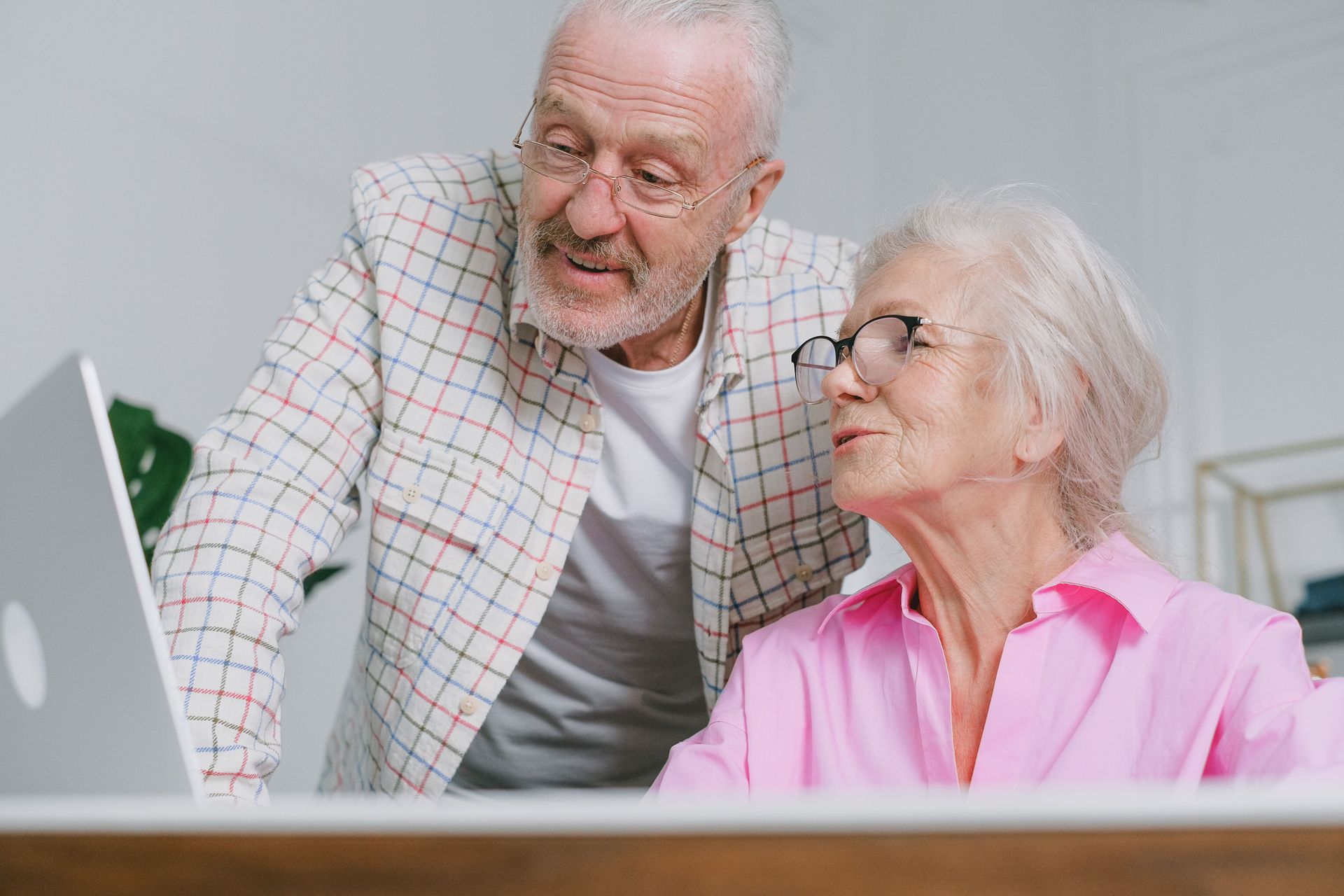Two people smiling at a laptop in a bright room, with an older white-haired man in a plaid jacket and a woman in pink.