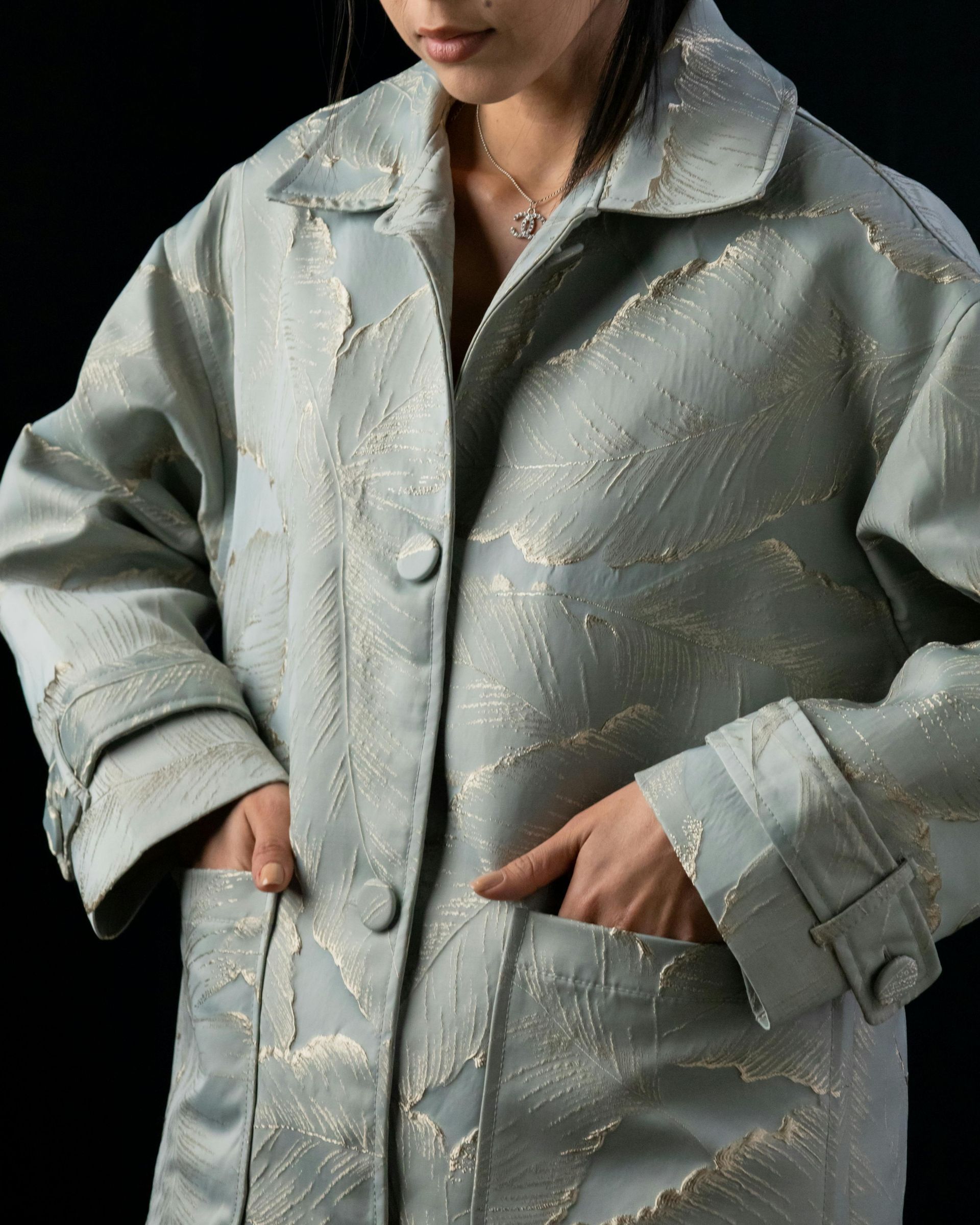 Woman in a long, light blue coat with gold embellishments, hands in pockets, against a black background.