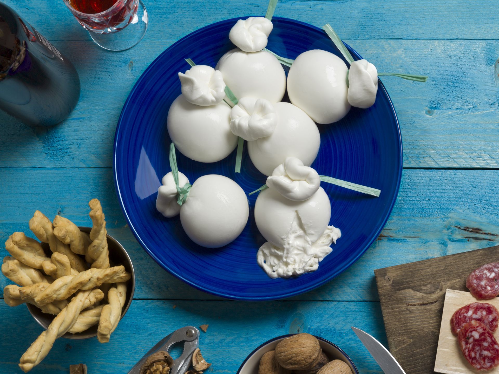 Burrata bianca su un piatto blu, con grissini, salame, noci e vino su un tavolo blu.