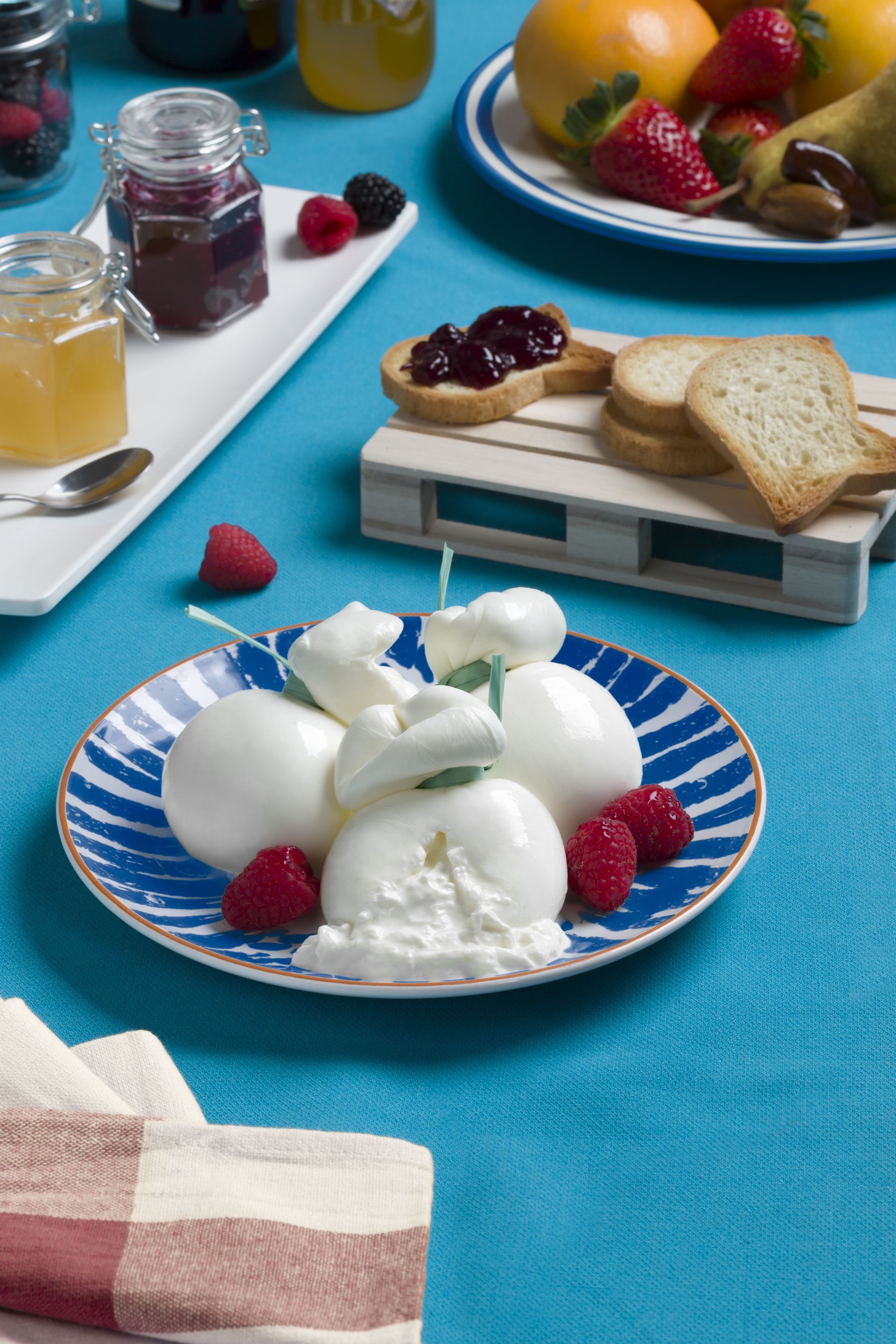 Burrata con lamponi su un piatto a strisce blu, accanto a marmellata, frutta e pane tostato, il tutto adagiato su una superficie blu.