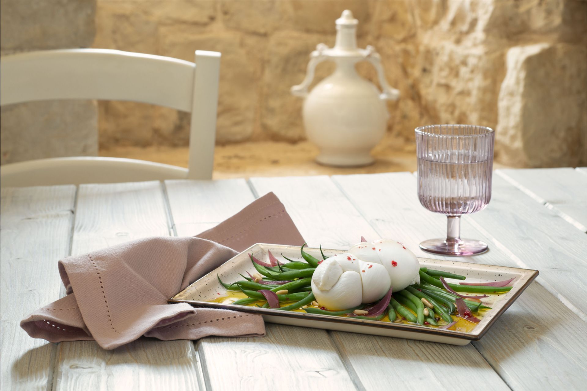Burrata con fagiolini e cipolle su un piatto rettangolare bianco. Sono presenti anche un bicchiere d'acqua e un tovagliolo rosa.