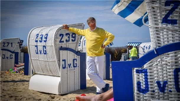 Ein Mann in einem gelben Pullover steht neben einem Korbstuhl am Strand.