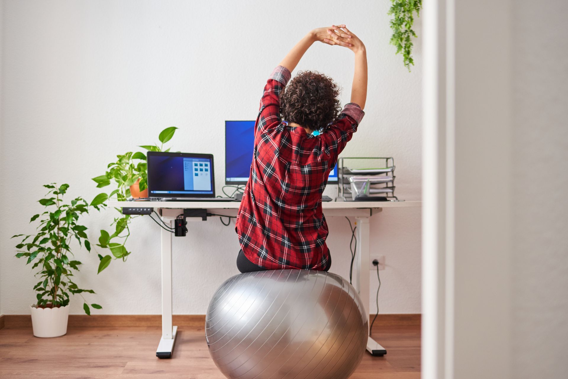 A woman is sitting on an exercise ball, stretching in front of a desk.