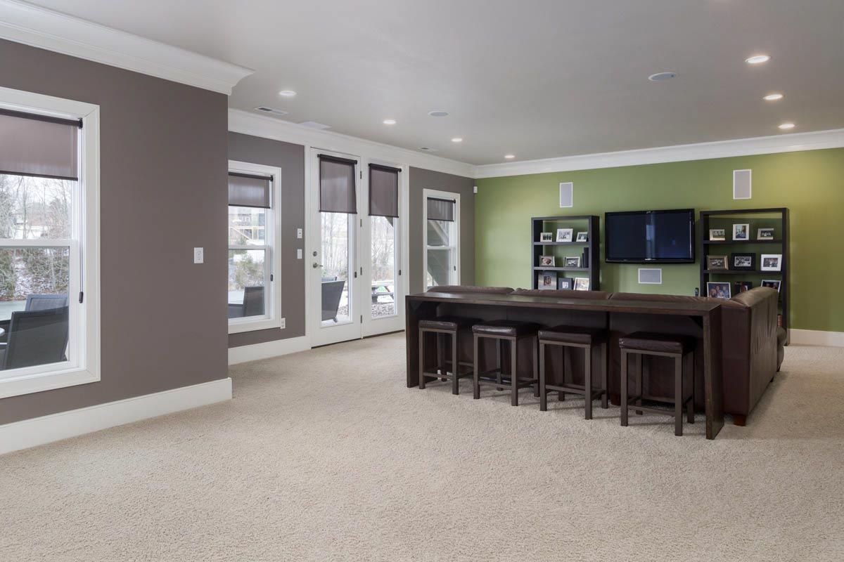 Spacious, carpeted room with built-in bar, TV, and seating. Features neutral colors, windows, and recessed lighting.
