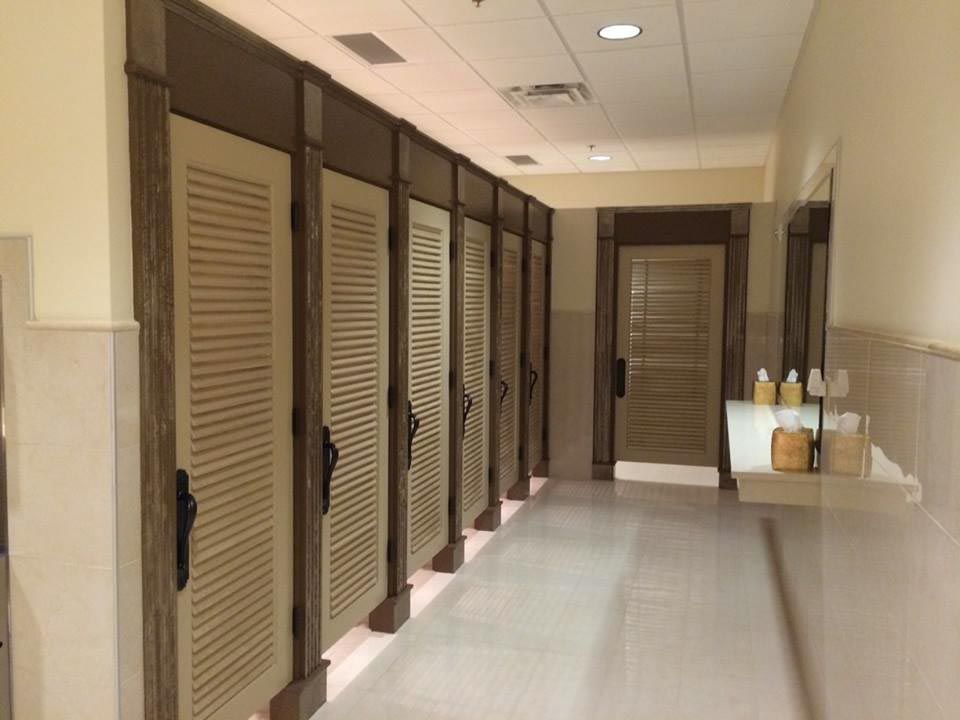Hallway with cream-colored restroom stalls with shuttered doors and a wall-mounted sink with toiletries.