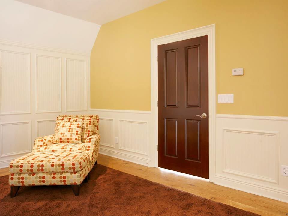 A room with a patterned armchair, brown door, and yellow walls with white molding.