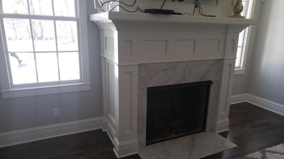 White fireplace with marble facing, flanked by windows, in a room with hardwood floors and gray walls.