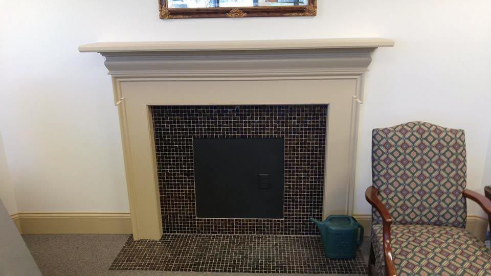 Fireplace with tile surround, black center, and ornate trim. A chair and watering can are nearby.