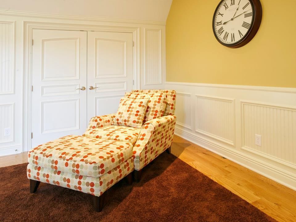 Chaise lounge with floral pattern on brown rug, against white paneling and double doors. A clock hangs on yellow wall.