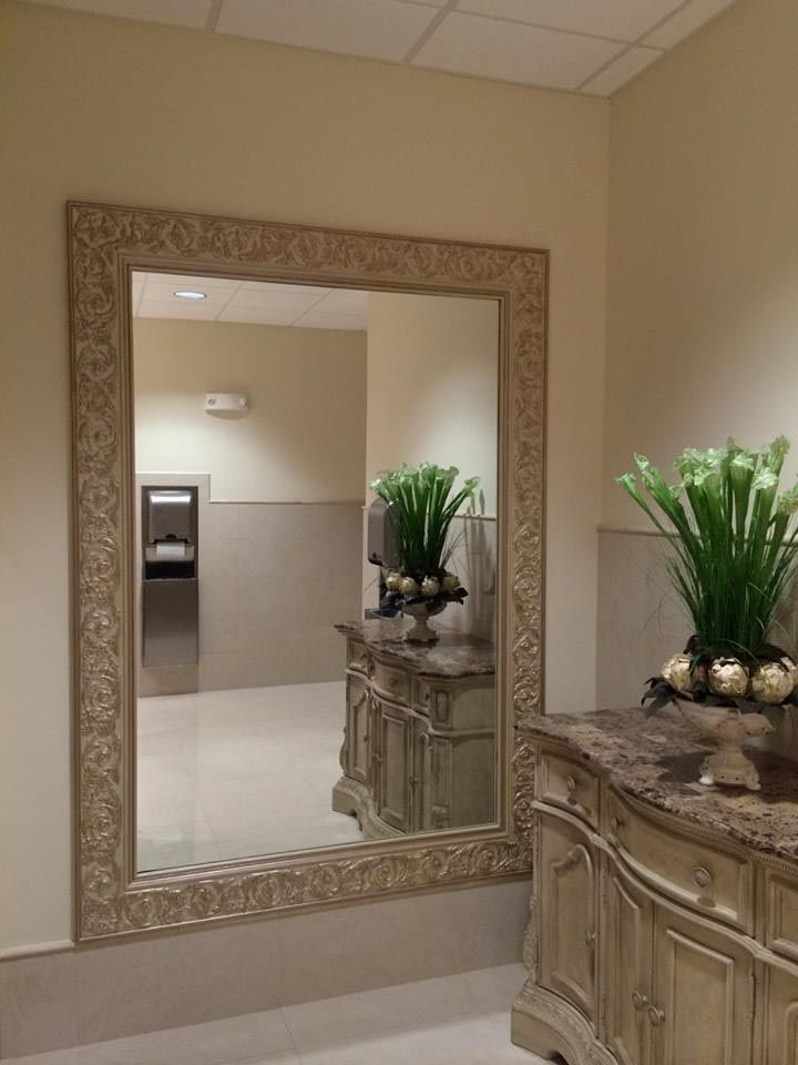 Large ornate mirror in a public restroom, reflecting the room's interior with a decorative countertop and flowers.