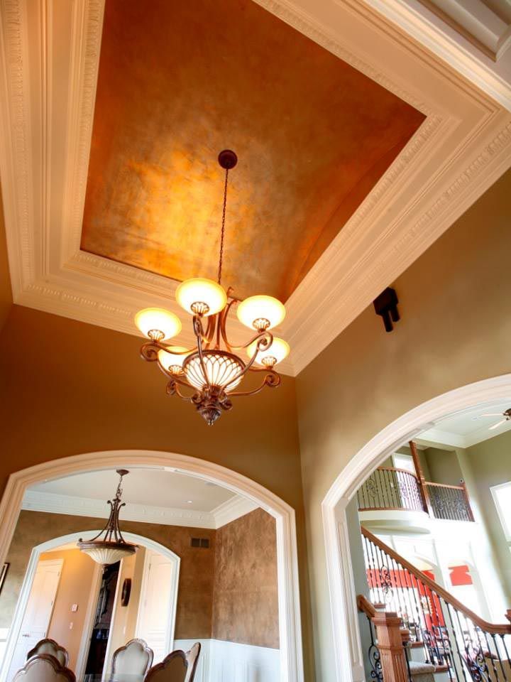 Ornate chandelier hangs from a decorative, metallic-painted ceiling, within a warmly colored room with arched doorways.