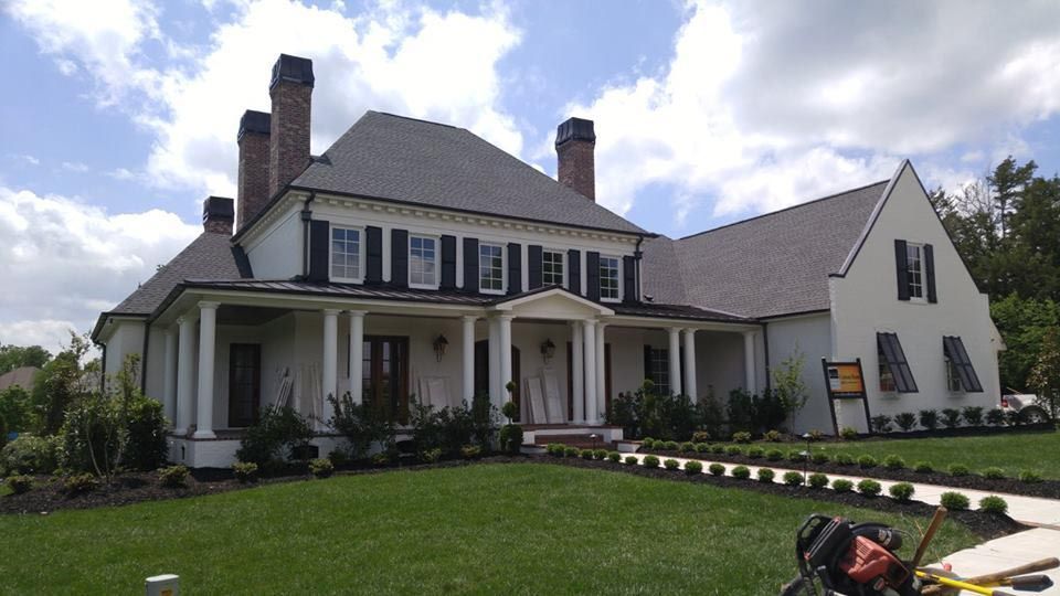 Large white house with black shutters, columns, and a dark roof. Green lawn with a row of shrubs.