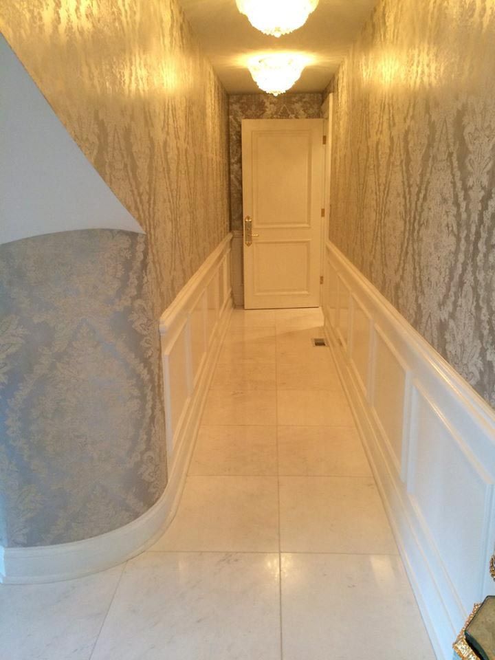 Narrow hallway with ornate wallpaper, white wainscoting, and a closed white door at the end.