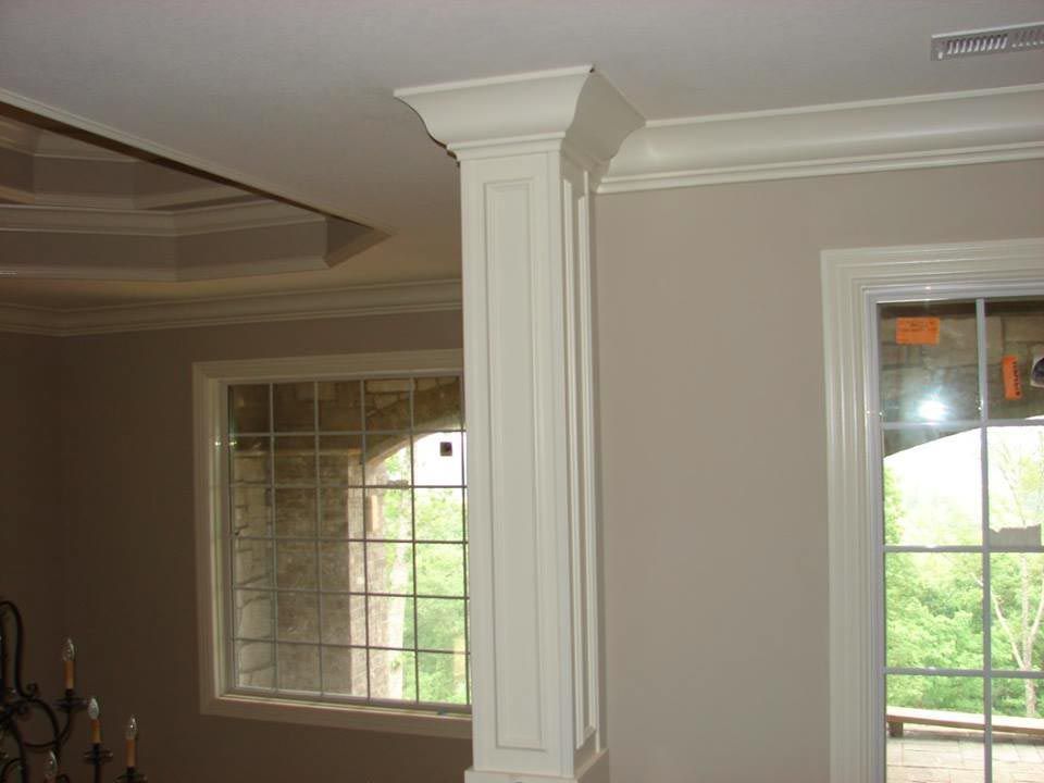 Cream-colored decorative column and crown molding in a room with light beige walls and large windows.