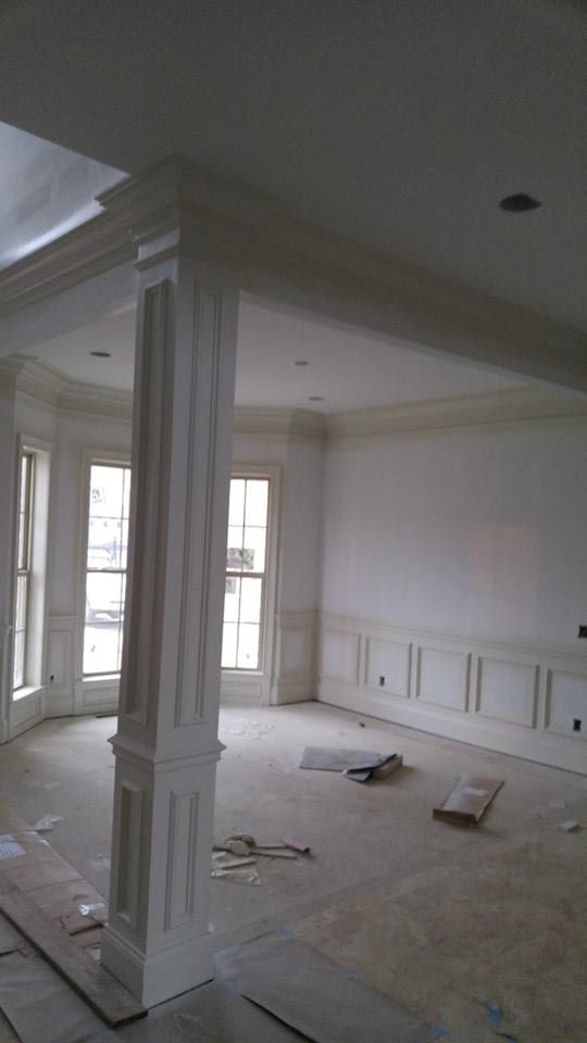 Interior of a room under construction with white pillars, crown molding, and wainscoting. Windows along the back wall.