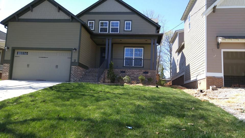 Green house with garage and porch, surrounded by grass and other buildings.