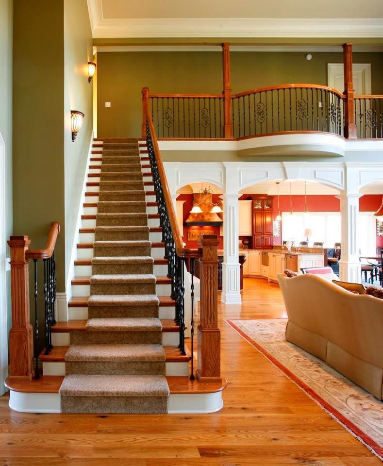 Spacious entry hall with staircase. Brown carpeted stairs, wooden railing, and green walls. View of kitchen and living room.