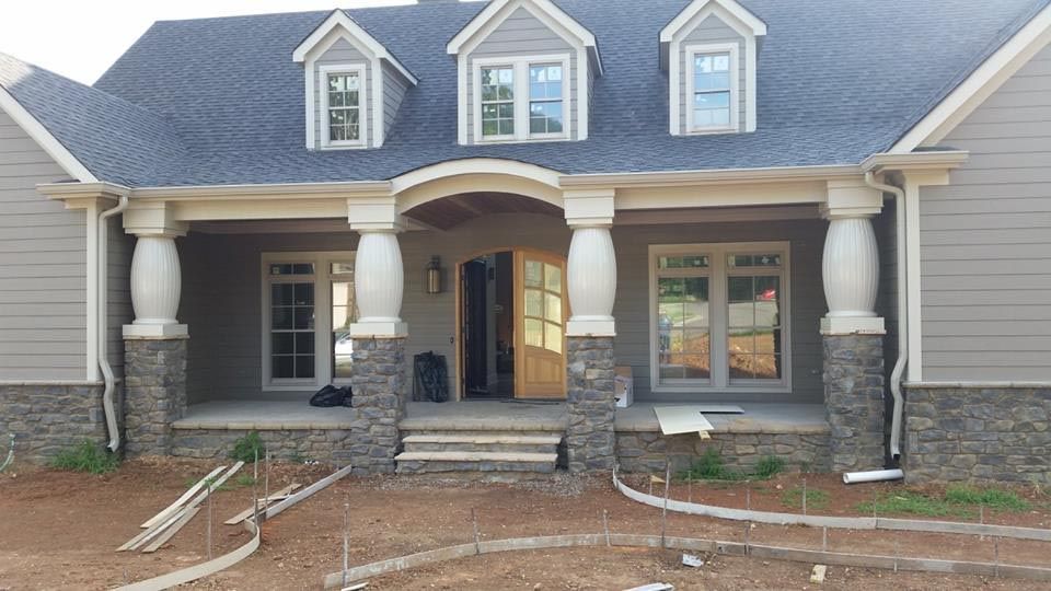 A light gray house with stone accents, a covered porch, and three dormer windows under construction.