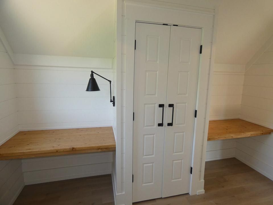 Built-in desks flank a closet with white paneled doors. Black wall-mounted lamp. Light wood countertops, white walls.