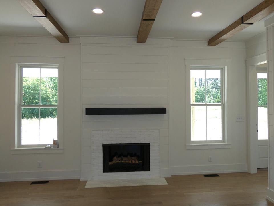 Living room with white walls, fireplace, and wood beams. Two windows flank the fireplace.