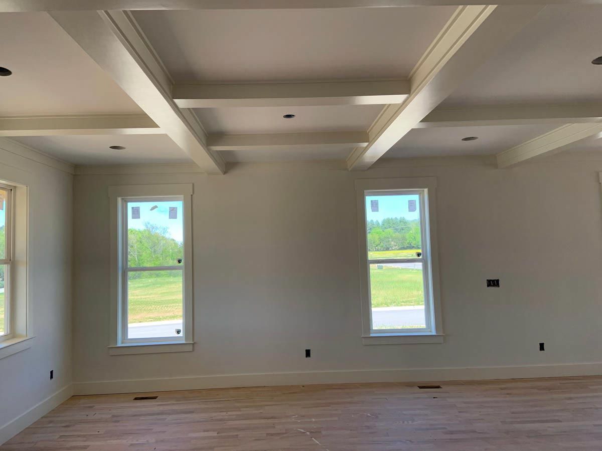 Empty room with white walls, exposed beams, windows, and light wood flooring.