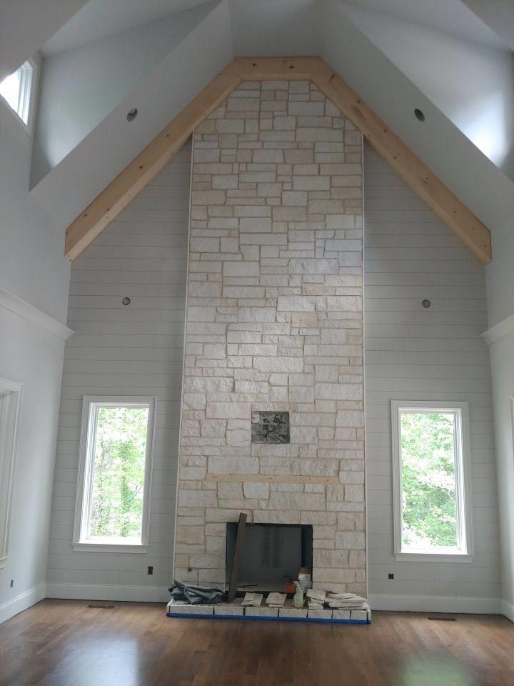 Stone fireplace with wood beams, flanked by windows, in a room with hardwood floors and high ceilings.