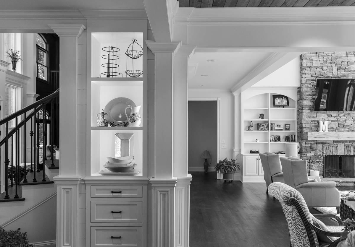 Interior view of a home's entryway. Built-in shelves, staircase, and open living area. Black and white.
